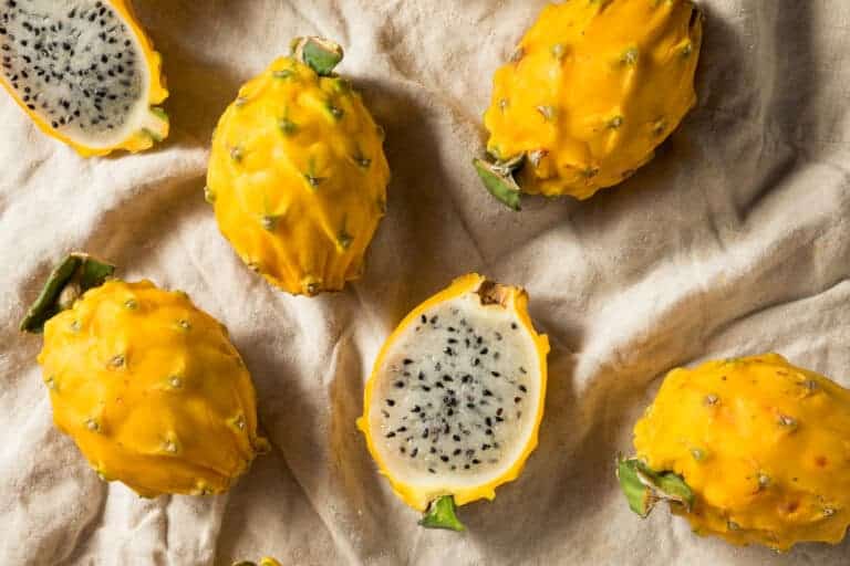 Exotic Colombian yellow dragon fruits are arranged on a beige cloth. One fruit is cut in half, revealing a white interior speckled with small black seeds. The bumpy skin features green-tipped protrusions, highlighting their unique appearance.