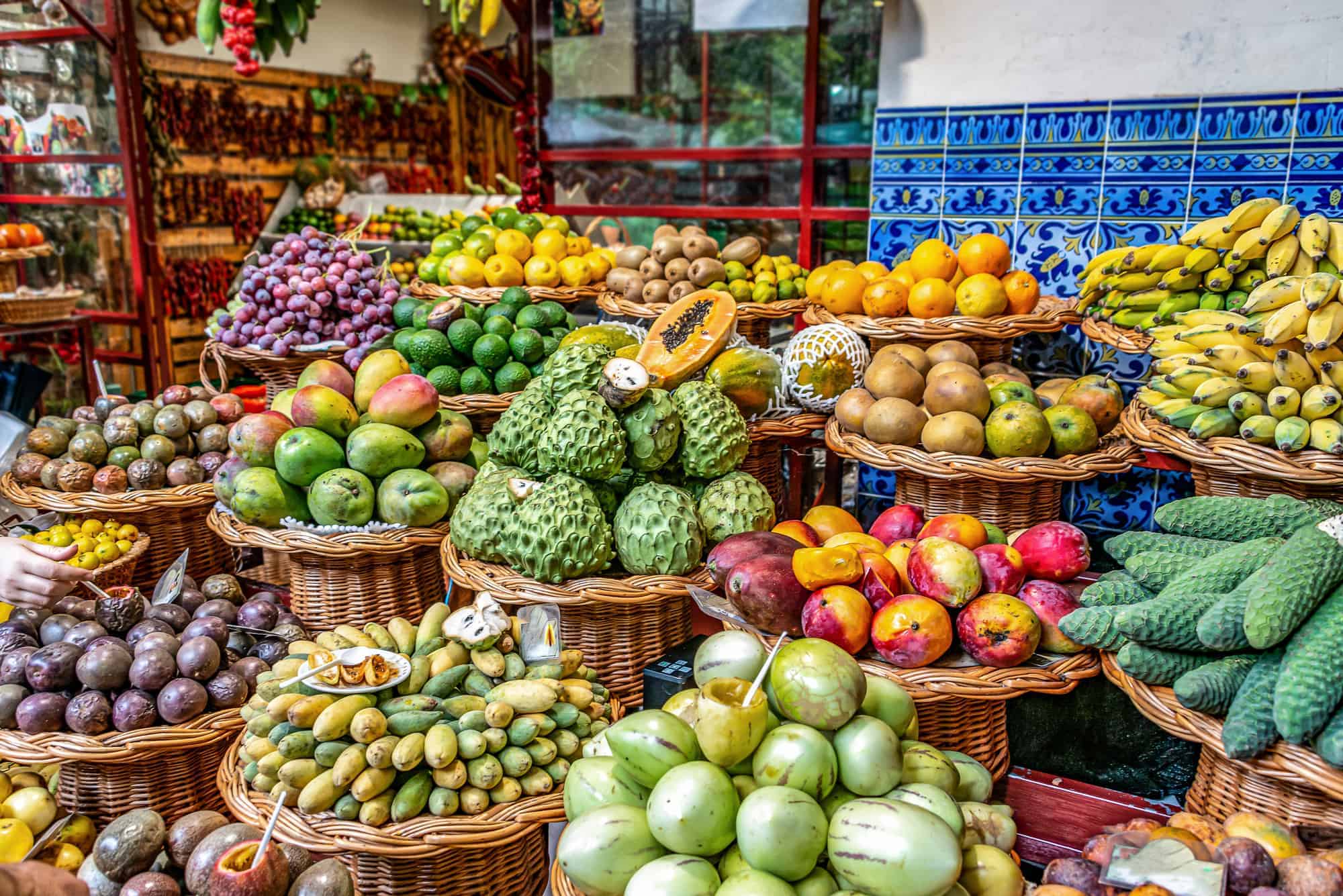 Baskets filled with a variety of exotic Colombian fruits, including grapes, mangos, passion fruit, bananas, and cherimoyas, are displayed at a market stall. The background features blue and white tiles and hanging dried peppers.