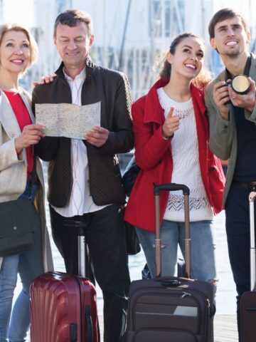 Four people stand together at a marina with sailboats. Two hold luggage, one holds a map, and another has a camera. They are casually dressed and appear to be enjoying a sightseeing moment by the water.
