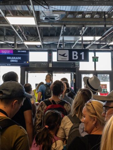 Passengers wait in line at an airport gate with signs for a flight to Belgrade. The area is crowded, and the ceiling shows exposed infrastructure. Travelers carry backpacks and luggage. A few gate monitors display flight details.
