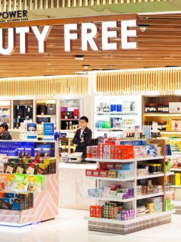 A brightly lit duty-free store in an airport features shelves stocked with snacks, drinks, and various products. A sign reads "DUTY FREE" above the entrance. Shoppers and staff are visible in the background, with items neatly organized.