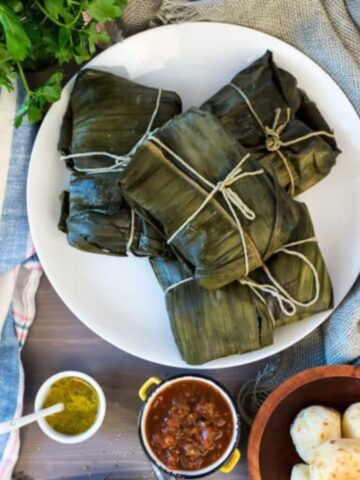 A white plate with four bundles wrapped in green leaves, tied with string, reminiscent of Colombian street food, sits on a table. Surrounding it are small bowls containing sauces, round breads, and a bunch of fresh parsley. A blue and white cloth is partly visible under the plate.