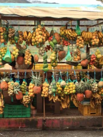 A market stall displaying a variety of hanging tropical and exotic Colombian fruits, including bananas, pineapples, and melons. Green crates are stacked under the wooden counter, and the scene is set outdoors with a canopy overhead.
