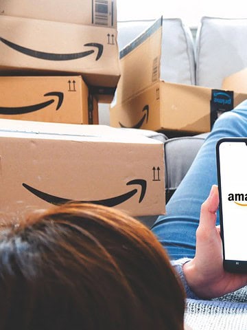 Person lying on a couch surrounded by Amazon boxes, holding a smartphone displaying the Amazon Prime logo. They are wearing blue jeans and the boxes are various sizes, all with the Amazon smile logo.