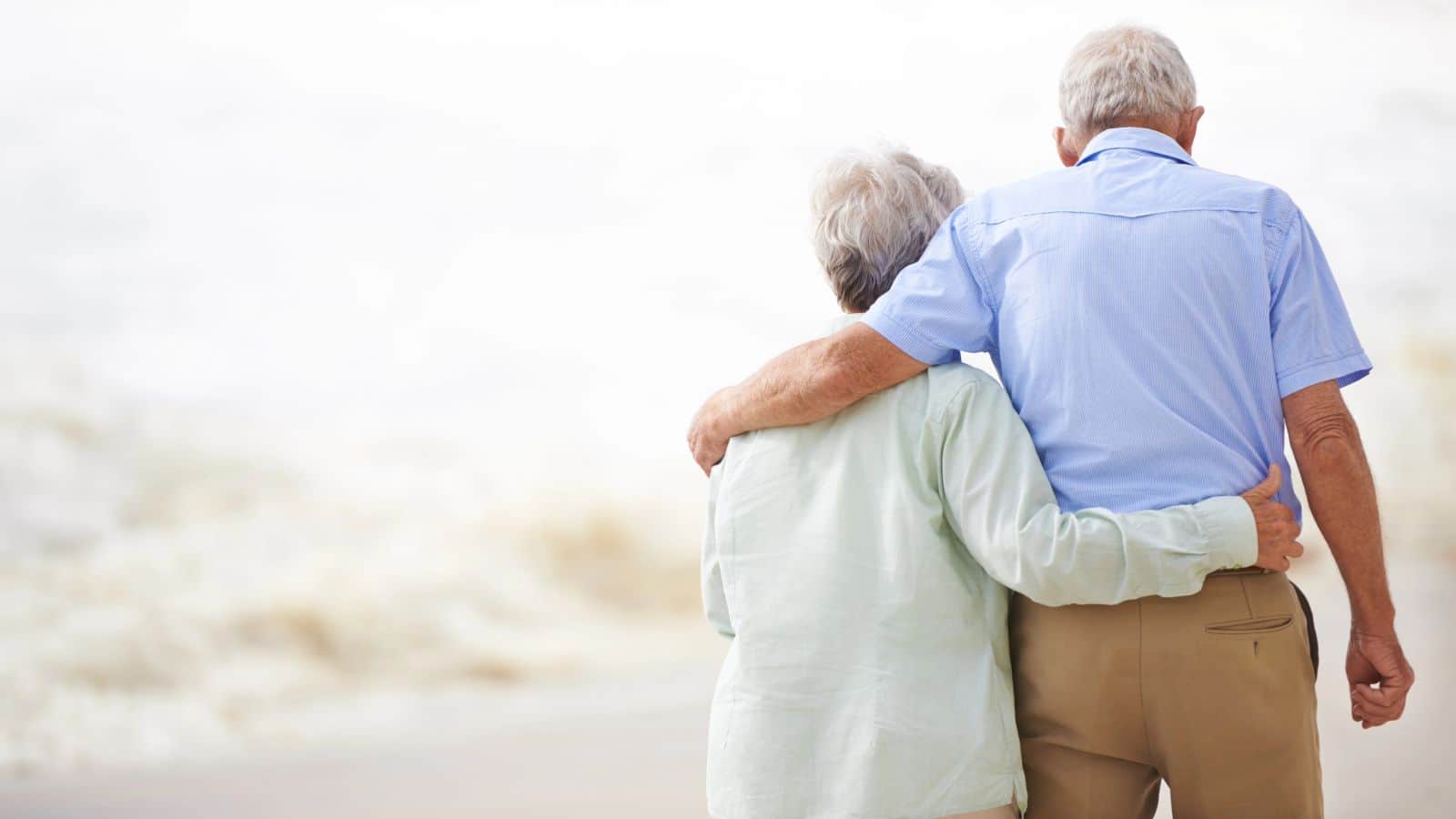 An elderly couple is walking on a beach with their backs to the camera. The person on the left has short gray hair and a light green shirt, while the person on the right has short hair and is wearing a light blue shirt and brown pants.