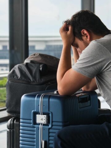 A person sits in an airport terminal, leaning forward with their head in their hands. They are wearing a gray shirt and sitting next to two suitcases. The background shows airport buildings and a runway through large windows.