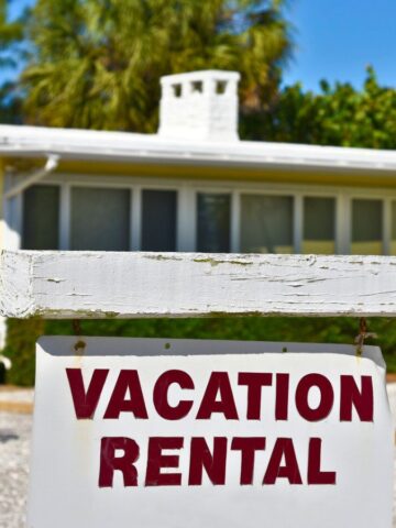 A white wooden sign with red text reads "Vacation Rental" in front of a yellow house surrounded by greenery and palm trees under a clear blue sky.
