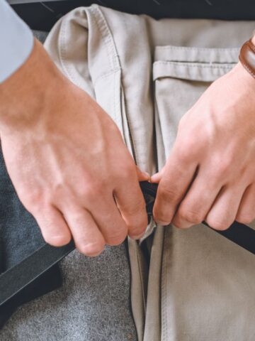 A person wearing a watch is packing a suitcase. The suitcase contains neatly folded clothing, including beige pants and gray fabric. The person is fastening a black strap over the clothes.