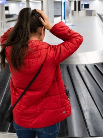 A woman in a red jacket stands at an empty airport baggage carousel with her hands on her head, facing away from the camera. The carousel is not moving, and the surrounding area appears quiet and empty.