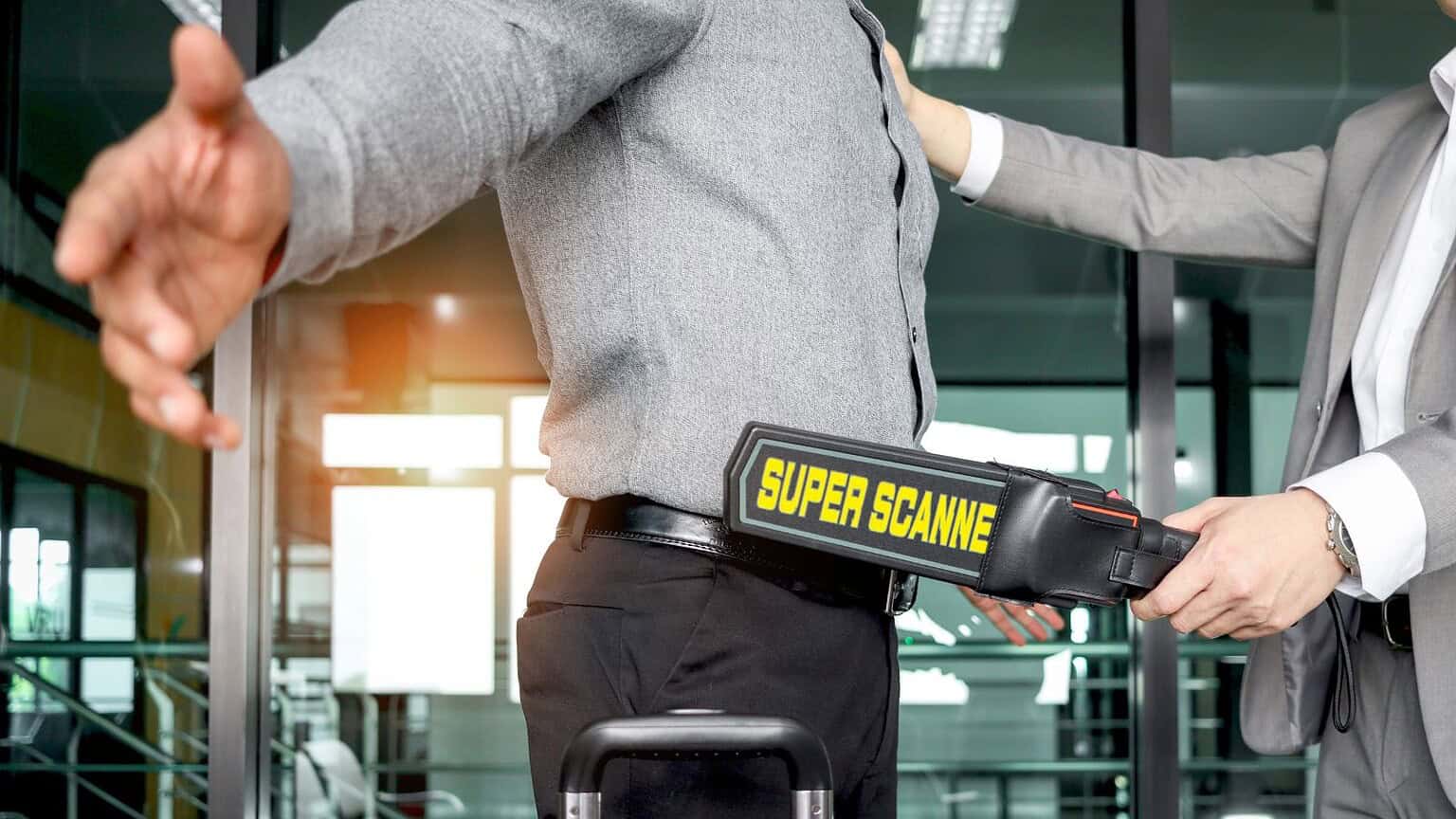 A security officer is scanning a man's body with a handheld metal detector labeled "Super Scanner" in an indoor setting. The man is standing with his arms stretched out while being checked.
