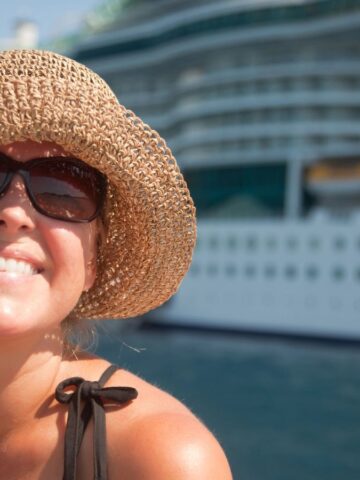 A woman wearing a straw hat and sunglasses smiles at the camera. In the background, a large cruise ship is docked in the water. The sky is clear and sunny.