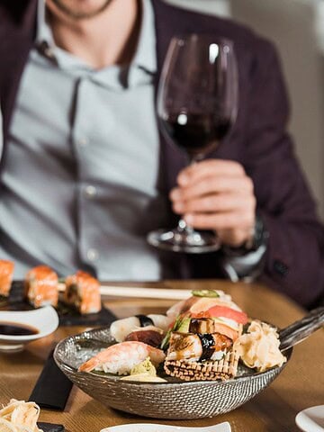A man and woman sit at a table with sushi rolls, sashimi, and dumplings on black slate plates. Both hold glasses of red wine. The woman wears a checkered shirt and the man is in a suit. Napkins and bowls of soy sauce are on the table.