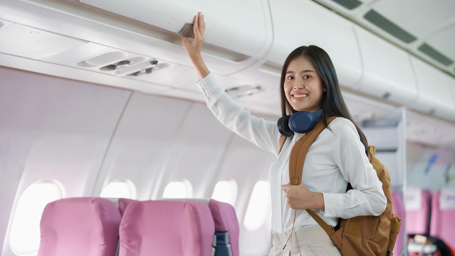 A woman in a white blouse and beige pants stands in an airplane aisle, smiling as she reaches for the overhead compartment. With headphones around her neck and a brown backpack on her shoulder, she’s careful to only take airplane items that belong to her. The seats are upholstered in pink fabric.