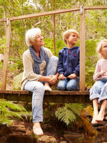 Two adults and two children sitting on a wooden bridge in a forested area. The adults, one wearing a light sweater and scarf, the other a blue sweater, are engaged in conversation with the children, who are dressed casually. Ferns and trees surround them.