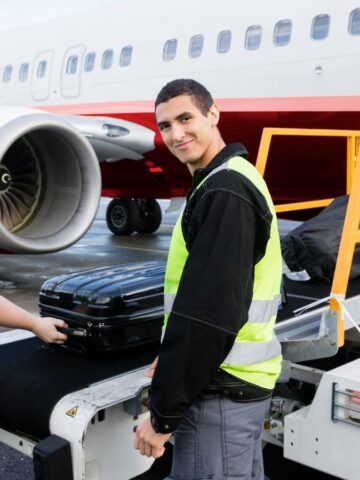 Two airport ground crew members, wearing yellow safety vests, load luggage onto a plane via a conveyor belt. One person smiles at the camera. An airplane is visible in the background.