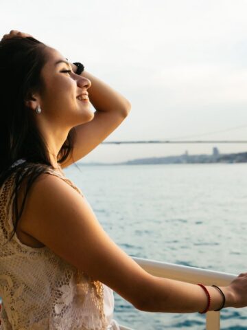 A woman with long hair stands on a boat, smiling and looking over the water. She rests one hand on the railing and the other on her head. A large bridge in the background spans across a wide body of water under a clear sky.
