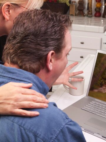 A couple sits in a kitchen, looking at a laptop showing an image of a cabin in the woods. The woman stands behind the man with her hand on his shoulder. A teapot and fruit are on the counter.