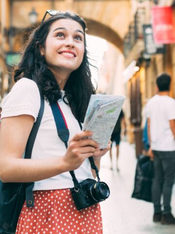 A woman in a white shirt and red polka dot skirt stands on a street holding a map. She has a camera around her neck and is wearing a backpack. The street is lined with old buildings, and there are a few people walking in the background.