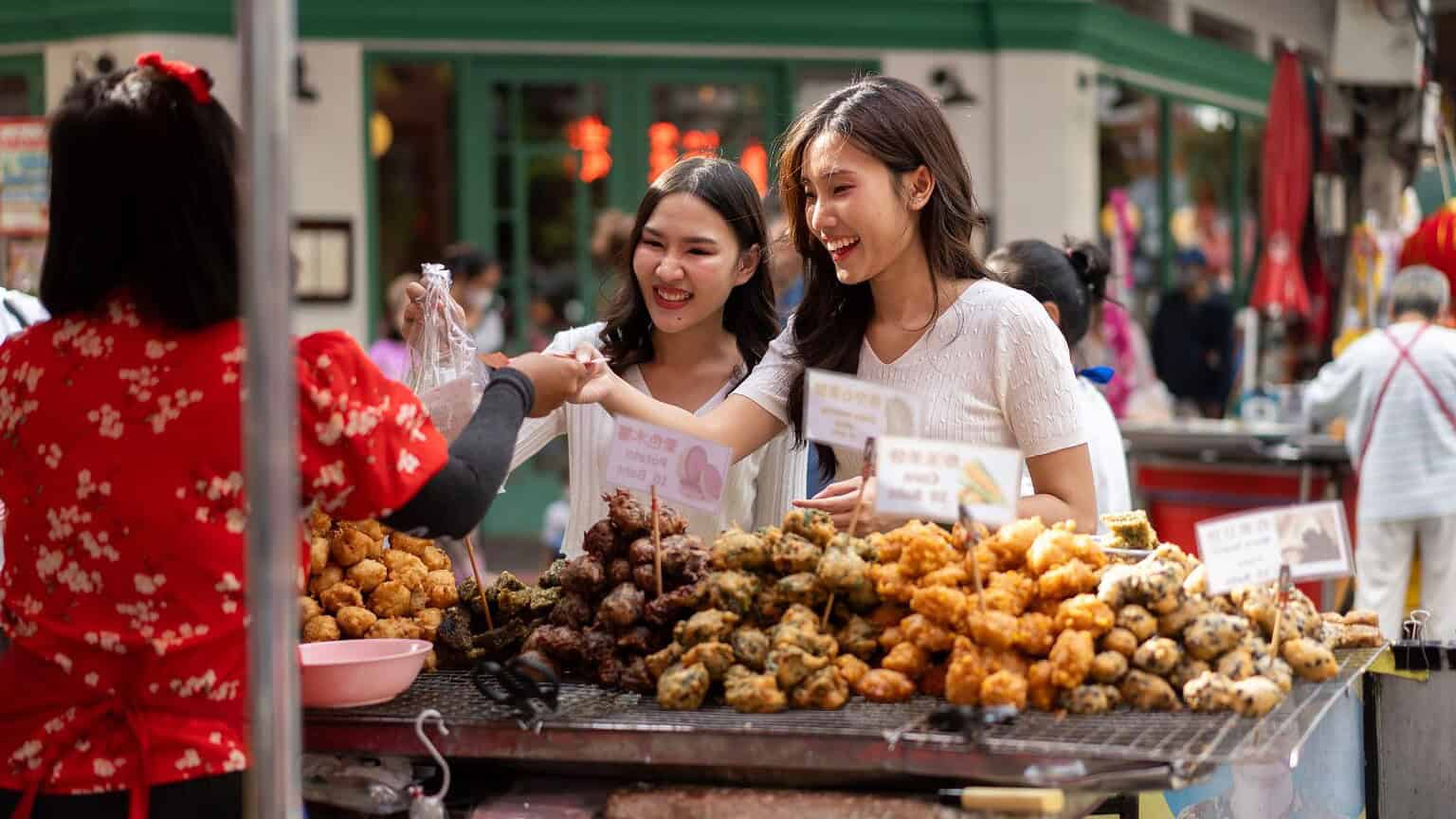Two women smile as they buy snacks from a street vendor. The vendor, wearing a red shirt, is handing them a bag of fried food from a tray filled with various items. People are walking in the background near a green storefront.