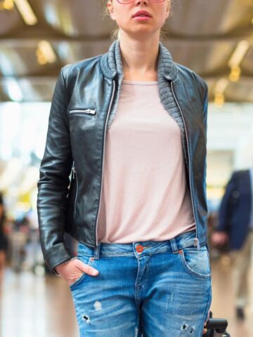 A person in a black leather jacket and blue jeans stands in a busy airport terminal. They are facing forward with hands in pockets. The background shows various travelers walking and signs indicating directions.