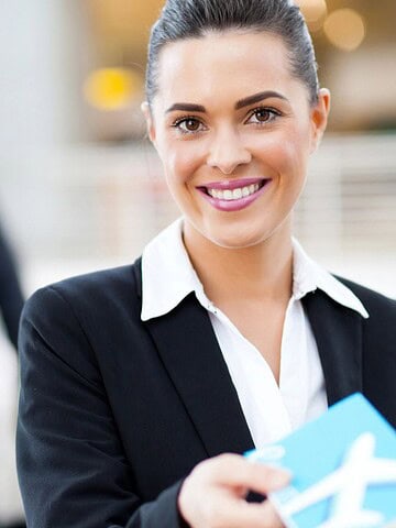 A woman in a business suit is smiling while holding a passport and boarding pass. She stands in focus in the foreground. A man in a suit is blurred in the background, looking at his phone. The setting appears to be an airport.
