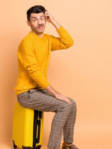 A man wearing a yellow sweater and plaid pants sits on a yellow suitcase against an orange background. He is scratching his head and has a surprised expression.