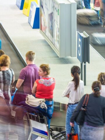 People stand and wait around a luggage carousel at an airport baggage claim. Some individuals have luggage carts, and the area appears busy and crowded. A sign marked with the number 5 is visible near the carousel.