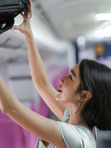 A woman with shoulder-length hair places a black backpack in the airplane's overhead compartment. She is wearing a smartwatch and standing in an aisle with empty pink seats visible in the background.
