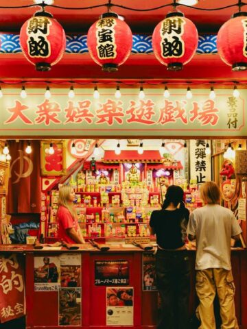 A vibrant street stall decorated with red paper lanterns and banners featuring Japanese text. Two people stand at the counter, one reading a menu. The booth has shelves displaying various colorful items and a red roof adorned with signs.