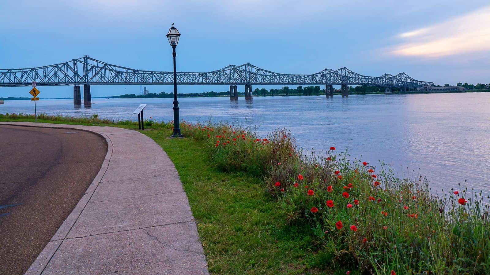 A wide river is spanned by a large steel truss bridge. A curved pathway lined with grass and red flowers runs along the riverbank. A lamppost stands by the path, and the sky is overcast with subtle hues of evening light.
