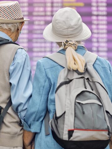 An elderly couple wearing hats and backpacks stands in an airport terminal, looking at a large purple-colored flight information display board.