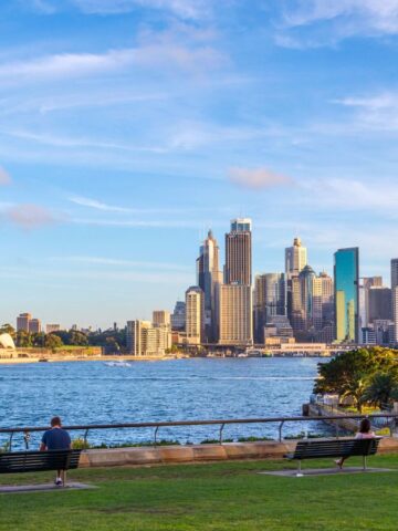 View of Sydney Harbour with the Opera House on the left and the city skyline in the background. A few people sit on benches in a park in the foreground. The sky is partially cloudy with a warm afternoon light.
