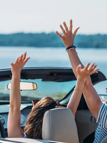 A couple is sitting in a convertible car with the top down by a body of water. Both have their hands raised in the air. The scene is daytime with a clear view of the water and distant shoreline.