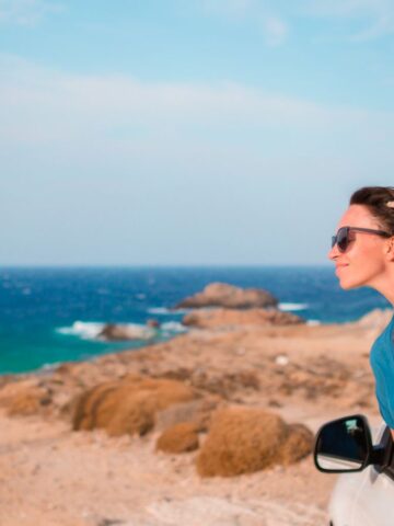 A person wearing sunglasses leans out of a car window, holding their hair in the wind. The background features a rocky coastline with the sea and a partly cloudy sky.