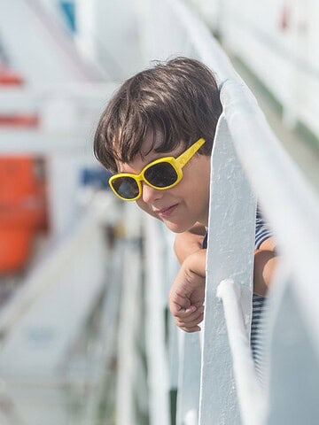 A child wearing yellow sunglasses leans over the railing of a boat, looking out at the water. An orange lifeboat is visible in the background. The scene is set against a bright blue sea.