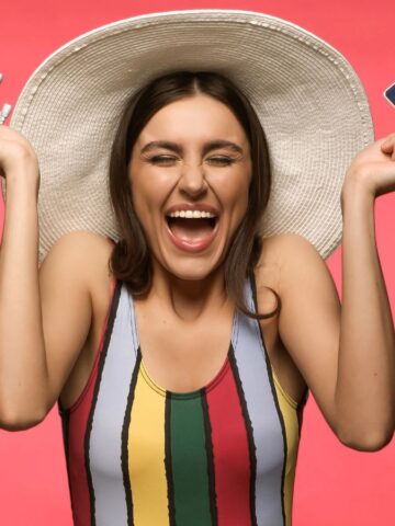 A woman wearing a wide-brimmed hat and a striped sleeveless top smiles widely, holding a toy airplane in one hand and a passport with a boarding pass in the other, standing in front of a pink background.