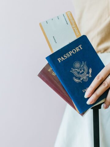 A person holds a blue U.S. passport, a boarding pass, and another passport while resting their hand on the handle of a suitcase. The person is wearing a light-colored outfit.