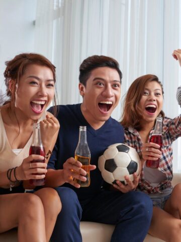 Six young adults sit on a couch, smiling and cheering while watching something off-camera. One is holding a soccer ball, and several are holding bottles of drinks. They appear to be enjoying a sports event together indoors.