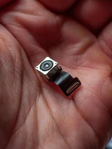 A close-up of a person's hand holding a small electronic component, specifically a camera module with a lens and ribbon cable connector.