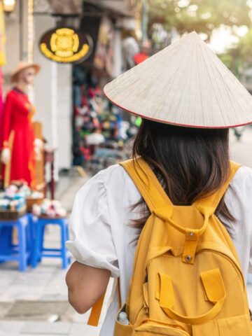 A person wearing a conical hat and yellow backpack walks along a busy street lined with shops selling hats and souvenirs. Motorbikes and people are visible in the background.