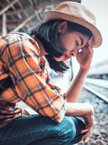 A person wearing a plaid shirt, jeans, and a hat sits on their haunches at a train station platform, holding their head with one hand, with a backpack on their back and train tracks beside them.