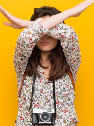 A person stands against a yellow background, arms crossed in an X above their face, wearing a butterfly-patterned shirt and a camera around their neck. Their face is partially covered by their arms.