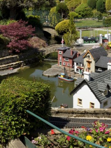 A young girl with long blonde hair in a striped dress looks at a detailed miniature village with small houses, water features, greenery, and flowers on a sunny day.