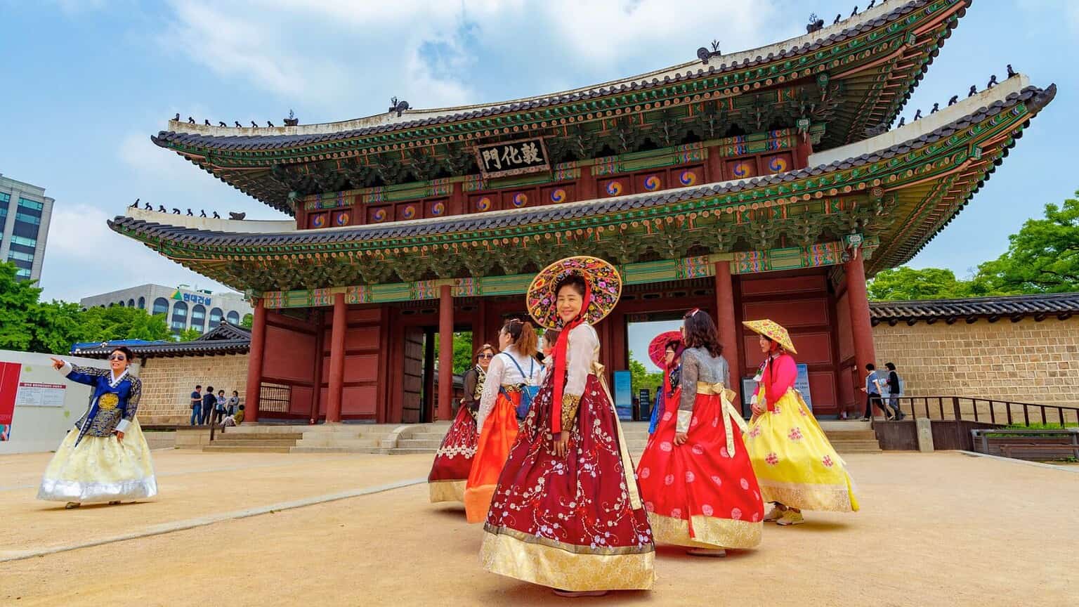 A group of people wearing colorful traditional Korean hanbok stand and walk in front of a large, ornate wooden palace gate with Korean writing, under a partly cloudy sky.