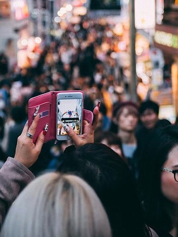 A crowded city street filled with people, some taking photos with smartphones. Bright lights and signs from shops and buildings line both sides of the street. The overall scene is busy and bustling.