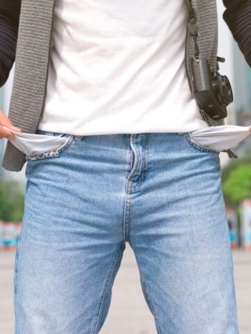 A person standing outdoors in jeans and a white t-shirt turns out both empty front pockets, revealing that they have no money. Buildings and trees are visible in the background.