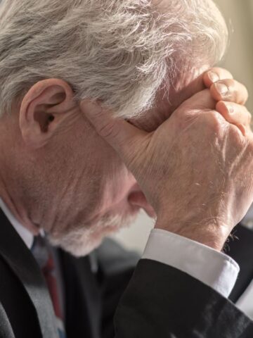An elderly man in a suit sits at a table, resting his forehead in his hands, appearing stressed or deep in thought. The background is blurred and a small plant is visible on the right side.