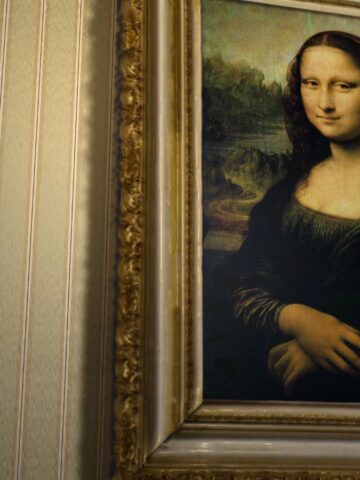 A framed painting of the Mona Lisa hangs on a patterned wallpapered wall. The portrait shows a woman with a neutral expression, dark hair, and folded hands, set against a distant landscape background.