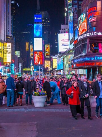 Crowd of people walking in Times Square, New York City, at night, surrounded by bright billboards and neon advertisements. The American flag is displayed on an electronic screen to the left.