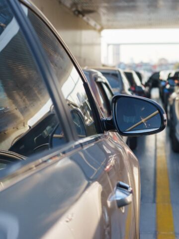 Cars are lined up in multiple lanes of heavy traffic, closely packed together under a covered area, with side mirrors and windows reflecting sunlight.